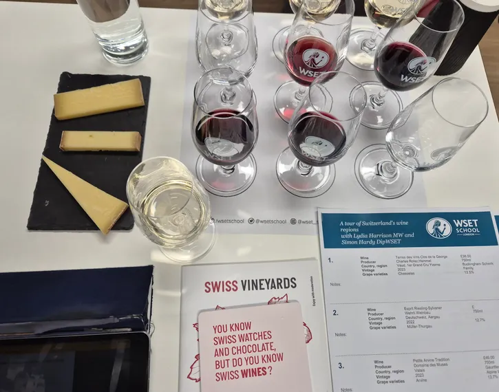 White table with wine tasting glasses filled with white and red wine, 3 pieces of cheese, a booklet about "Swiss Vineyards" & a WSET School tasting sheet with the wine list.